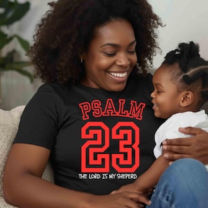 Peut inclure: T-shirt noir avec le texte "PSALM 23" en lettres rouges. En dessous du chiffre 23 se trouve le texte "THE LORD IS MY SHEPERD" en rouge. Le t-shirt est porté par une personne.