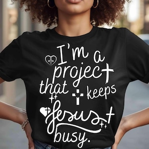 Peut inclure: T-shirt noir avec l'inscription blanche "I'm a project that keeps Jesus busy". Le motif comprend un cœur avec une croix, une croix et des étoiles. Le t-shirt est de style col rond.
