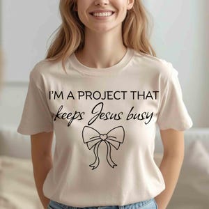 Peut inclure: T-shirt crème avec le texte noir "I'M A PROJECT THAT keeps Jesus busy". Un motif de nœud noir est centré sous le texte. Le t-shirt a un col rond et des manches courtes.