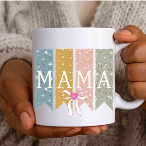 Peut inclure: Tasse en céramique blanche avec le mot "MAMA" en lettres majuscules blanches. Le mot est placé sur quatre bannières verticales de couleurs pastel, chacune avec un motif floral. Un nœud rose avec un cœur est en dessous.