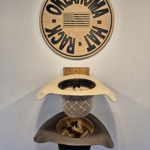 May include: Wooden "Oklahoma Hat Rack" sign with an American flag, above two cowboy hats. The hats are a light tan and gray, with black and gold interiors. The sign is circular and mounted on a white wall.