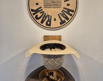 May include: Wooden "Oklahoma Hat Rack" sign with an American flag, above two cowboy hats. The hats are a light tan and gray, with black and gold interiors. The sign is circular and mounted on a white wall.