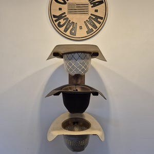 May include: A wooden hat rack with three levels, each designed to hold a hat. The rack is mounted on a white wall, with a circular sign at the top that reads "Oklahoma Hat Rack" and features an American flag.