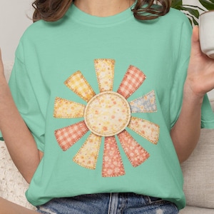 May include: A light green t-shirt featuring a colorful sunburst design. The sunburst is made of various patterned fabric pieces in shades of yellow, orange, and pink. The shirt is paired with denim shorts.