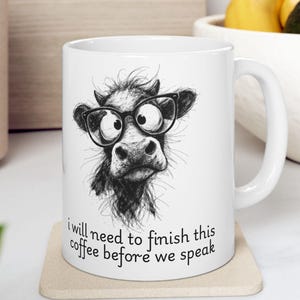 May include: White ceramic coffee mug featuring a whimsical black and white illustration of a cow wearing glasses. The mug has the text "I will need to finish this coffee before we speak".
