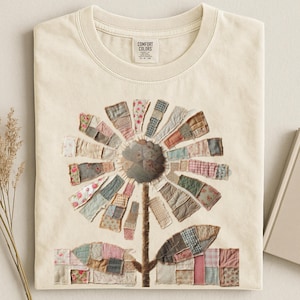 May include: Cream-colored t-shirt featuring a large, patchwork flower design. The flower's petals are made of various fabric patterns in muted tones. The shirt has a crew neck and a tag that says "Comfort Colors".