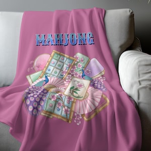 May include: A pink throw blanket with the word "MAHJONG" in blue lettering. The blanket features a design of peacock feathers, fans, and tiles in shades of pink, green, and purple. The blanket is draped over a gray sofa.