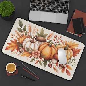 Puede incluir: Alfombrilla de ratón rectangular con diseño otoñal. La ilustración presenta calabazas, setas y hojas de otoño en tonos naranja, blanco y marrón. Un ratón de ordenador descansa sobre la alfombrilla, junto a un portátil y papelería.