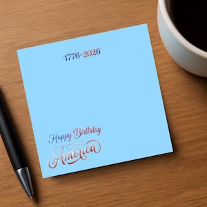 May include: A light blue square card with the text "1776-2026" at the top and "Happy Birthday America" in a decorative script at the bottom. A black pen and a white mug are partially visible.