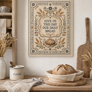 Könnte beinhalten: Ein gerahmtes Wandbild mit beigem Stoffhintergrund, blauen und braunen Blumenstickereien, Weizenähren und einer zentralen Brotillustration. Der Text lautet: GIVE US THIS DAY OUR DAILY BREAD Matthew 6:11 in dunkelblauer Schrift.