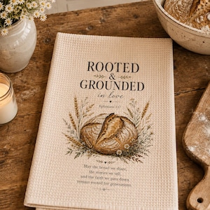 May include: A cream-colored waffle-weave kitchen towel featuring the black text "ROOTED & GROUNDED in love" above an illustration of a round loaf of bread surrounded by wheat stalks and small flowers, with the text "Ephesians 3:17" and a verse.