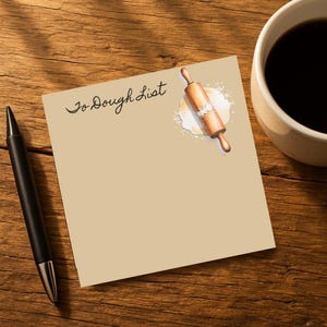 May include: A notepad with the words "To Dough List" in cursive, a rolling pin illustration, and a cup of coffee on a wooden table. A black pen is on the left side of the notepad.
