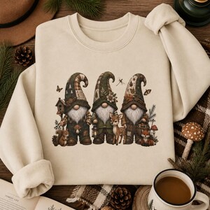 May include: A beige crewneck sweatshirt featuring a graphic print of three gnomes in forest-themed attire. The gnomes are surrounded by woodland elements including a squirrel, a deer, a fox, mushrooms, a birdhouse, and a lantern.