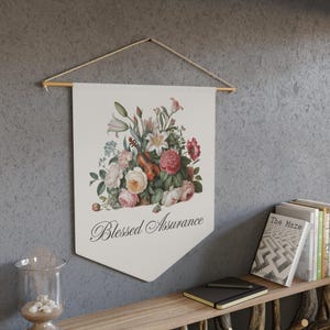May include: A cream-coloured wall banner featuring a floral arrangement and a violin. The arrangement includes pink, red, and white flowers. The banner has the words "Blessed Assurance" in script. It is hung with a wooden dowel and twine.