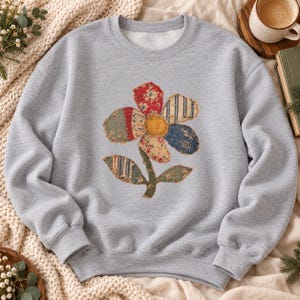 May include: A heather gray crewneck sweatshirt featuring a central graphic of a flower constructed from various fabric patches. The flower petals and leaves display diverse patterns including floral prints, stripes, and solid colors.