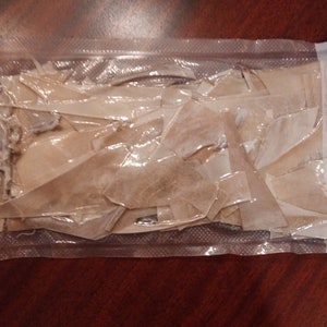 May include: A clear plastic bag containing numerous irregularly shaped, light brown pieces. The bag is sealed and appears to be vacuum-packed. The contents are tightly packed, with some pieces overlapping.