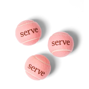 May include: Three pink tennis balls with the word "serve" printed in brown. The tennis balls have white seams and are set against a white background. The balls are a light shade of pink.