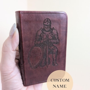 May include: A burgundy leather-bound book featuring an engraved image of a knight in armour, kneeling with a sword and shield. The shield has a cross design. The text "CUSTOM NAME" is visible on the bottom right.