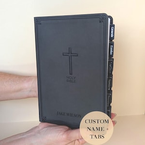 May include: A black leather Holy Bible with a cross embossed on the cover. The book has labeled tabs and the name "Jake Wilson" is printed on the bottom. The image also includes a circular sticker that says "CUSTOM NAME + TABS".
