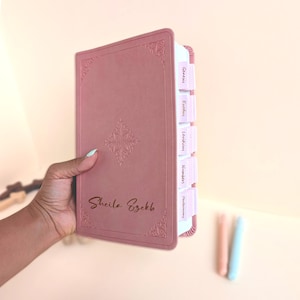 May include: A dusty rose-coloured Bible with a smooth cover featuring an embossed floral design in the centre and corners. The name Sheila Ezekb is printed in dark script on the front. It includes light pink index tabs labelled Genesis, Exodus, Leviticus, Numbers, and Deuteronomy.