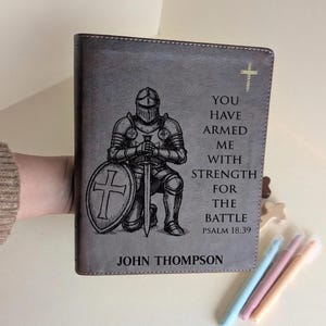 May include: Grey leather book cover with an engraved knight in armour holding a sword and shield with a cross. The text reads "YOU HAVE ARMED ME WITH STRENGTH FOR THE BATTLE PSALM 18:39" and "JOHN THOMPSON".