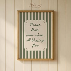 May include: A framed print with a gold-colored frame. The print features a cream-colored background with green vertical stripes. The center of the print has a cream-colored decorative shape with the words "Praise God, from whom all blessings flow" in a script font.