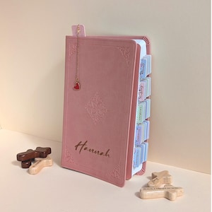 May include: A dusty pink Bible with a gold chain bookmark featuring a red heart charm. The cover is embossed with the name Hannah and decorative corner patterns. The pages include colourful index tabs labelled with book names like Genesis and Exodus.