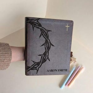 May include: Grey leather-bound book featuring a dark thorn design and a gold cross. The name "AARON SMITH" is printed at the bottom. Pastel pens and a wooden star are next to the book.