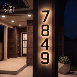 May include: A vertical, wall-mounted house number sign displaying the digits 7849 in a bold, black font. The numbers are backlit with a warm, glowing light, creating a halo effect against the rectangular, light-coloured mounting panel.