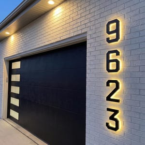 May include: Vertical house numbers 9, 6, 2, and 3 mounted on a white brick wall. The black metal numbers are backlit with a warm yellow glow, creating a halo effect against the textured brick surface next to a black garage door with windows.