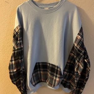 Może przedstawiać: Jasnoniebieska bluza z długimi rękawami i kieszenią w czarno-biało-czerwoną kratę. Bluza ma okrągły dekolt i luźny krój. Wzór w kratę to klasyczny design.