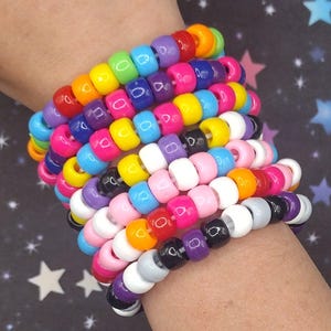May include: A stack of multiple bracelets made from colorful plastic pony beads in various shades including red, orange, yellow, green, blue, purple, pink, white, black, and gray, arranged in a repeating pattern on a clear elastic cord.