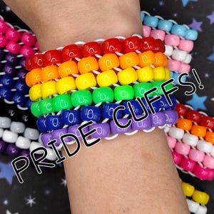 May include: A multi-row bracelet made of vibrant, glossy plastic pony beads arranged in a rainbow color sequence of red, orange, yellow, green, blue, and purple, strung together with white elastic cord. The text PRIDE CUFFS! is visible.