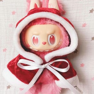 May include: A plush toy featuring a creature with pink fur and a red and white hooded cape tied with a white bow. The toy has a light skin tone, red eyes, and a small pink nose. The toy has a tag that says "E MONSTER".
