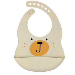 Puede incluir: Babero de silicona color crema para bebé con diseño de cara de oso. El babero presenta una nariz redonda naranja, ojos negros y pequeñas pecas. Tiene una correa ajustable para el cuello y un bolsillo curvo en la parte inferior.