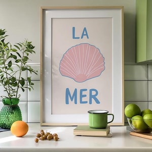 Könnte beinhalten: Ein gerahmter Kunstdruck mit einer hellrosa Muschelillustration, die zwischen dem blauen Text LA und MER auf einem hellen Hintergrund zentriert ist. Der Druck befindet sich in einem hellen Holzrahmen und zeigt ein klares Grafikdesign.