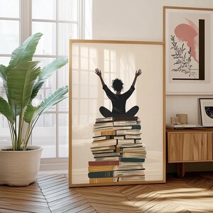 Könnte beinhalten: Ein gerahmter Wandkunstdruck, der die Silhouette einer Person mit erhobenen Armen zeigt, die auf einem hohen Stapel Bücher sitzt. Die Illustration verwendet eine neutrale Farbpalette mit Schwarz, Creme und erdigen Tönen in einem hellen Holzrahmen.
