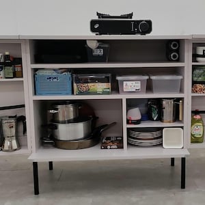 Könnte beinhalten: Ein weißer Schrank mit mehreren Regalen, schwarzen Beinen und einem schwarzen Gerät oben. Die Regale sind mit verschiedenen Küchenartikeln gefüllt, darunter Töpfe, Teller, Lebensmittelbehälter und Kaffeemaschinen. Schwarze Riemen sichern die Gegenstände.