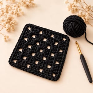 May include: A black square crochet granny square motif made from thick yarn, accompanied by a matching ball of black yarn and a gold-colored metal crochet hook with a black handle, arranged on a light beige surface.