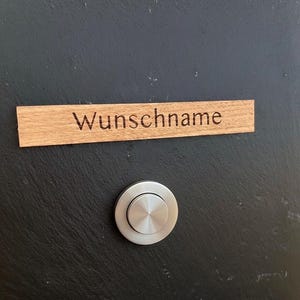 May include: A wooden nameplate with the word "Wunschname" engraved on it, mounted above a circular, brushed metal doorbell button. The nameplate is rectangular and made of light-colored wood. The doorbell button is silver and has a circular design.