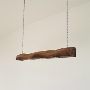 Può includere: Un lampadario rustico in legno a forma di trave, sospeso al soffitto con due catene argentate. La trave ha una forma naturale e ondulata e un ricco colore marrone. L'apparecchio è progettato per l'illuminazione ambientale.