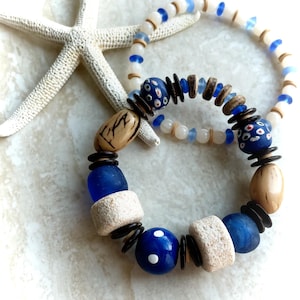 Könnte beinhalten: Zwei Armbänder mit blauen, weißen und braunen Perlen. Ein Armband hat große blaue Perlen mit weißen Punkten, das andere kleinere blaue und weiße Perlen. Im Hintergrund befindet sich ein weißer Seestern.