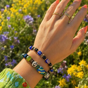 Könnte beinhalten: Zwei Perlenarmbänder mit einer Mischung aus blauen, grünen, schwarzen und weißen gemusterten Perlen, einschließlich floraler und abstrakter Designs, getragen mit zwei goldfarbenen Metallringen an den Fingern, einer mit strukturiertem Band und einer mit Lochmuster.