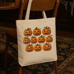 May include: A cream-colored tote bag featuring a repeating pattern of orange jack-o'-lanterns with carved faces and green stems. The bag is made of a sturdy fabric and has long handles for carrying over the shoulder.