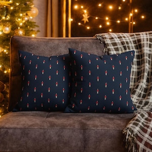 Może przedstawiać: Dwie granatowe poduszki z powtarzającym się wzorem czerwonych i złotych figurek świec. Poduszki leżą na brązowej sofie z pledem w kratę. W tle choinka i łańcuchy świetlne.