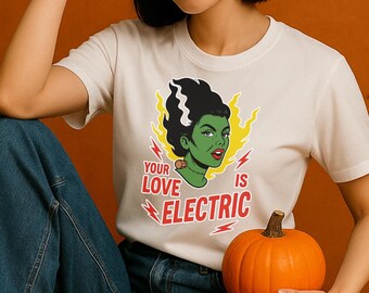 May include: Off-white t-shirt featuring a graphic of a green-skinned woman with black hair, red lips, and the text "YOUR LOVE IS ELECTRIC." The design includes yellow lightning bolts and red text. An orange pumpkin is held in the foreground.