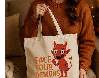 May include: A beige tote bag featuring a cartoon devil with the text "FACE YOUR DEMONS" in orange. The devil is red with horns, a tail, and a friendly expression. The bag has long, beige handles.