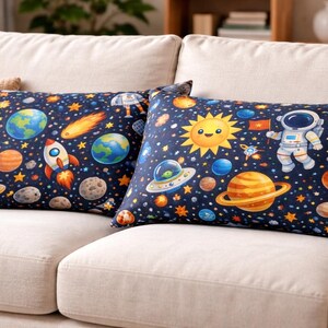 Op de afbeelding: Twee rechthoekige kussens met een marineblauwe achtergrond met een kleurrijk ruimtethema-ontwerp. Planeten, sterren, raketten en een lachende zon zijn afgebeeld. Eén kussen toont een astronaut. De kussens liggen op een crèmekleurige bank.