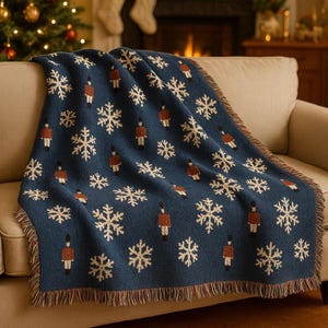 May include: A navy blue throw blanket with a repeating pattern of snowflakes and toy soldier figures. The blanket has a fringed edge and is draped over a beige sofa. The background includes a Christmas tree and a fireplace.
