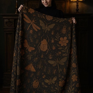 Può includere: Una coperta marrone scuro con un motivo ripetuto di insetti e fiori in un colore marrone più chiaro. La coperta presenta libellule, farfalle, coleotteri ed elementi floreali. La coperta è tenuta da una persona.